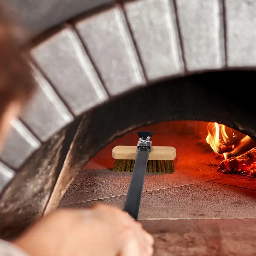 Brass pizza oven cleaning brush sweeping the floor of a lit wood-fired oven